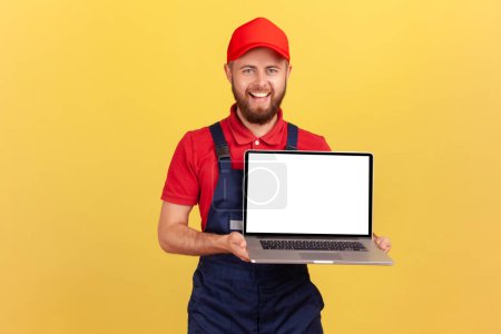 Photo for Portrait of smiling happy worker man in uniform standing and showing laptop with blank screen for advertisement, looking smiling at camera. Indoor studio shot isolated on yellow background. - Royalty Free Image