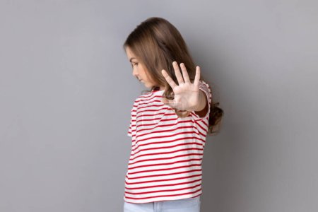 Photo for Portrait of scared serious little dark haired girl wearing striped T-shirt showing block stop gesture, set back bullying and violence. Indoor studio shot isolated on gray background. - Royalty Free Image