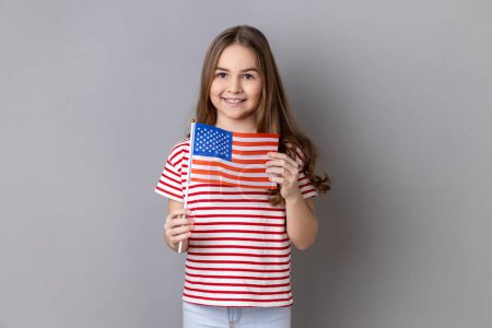 Photo for American flag. Portrait of delighted happy adorable cute little girl wearing striped T-shirt holding flag of United States of America, looking at camera. Indoor studio shot isolated on gray background - Royalty Free Image