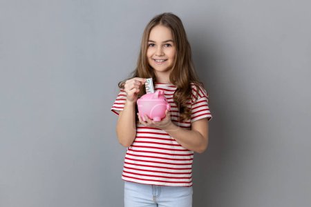 Foto de Retrato de una adorable niña sonriente con una camiseta a rayas de pie sosteniendo la colocación de billetes en una alcancía rosa en las manos, ahorrando dinero. Estudio interior plano aislado sobre fondo gris. - Imagen libre de derechos