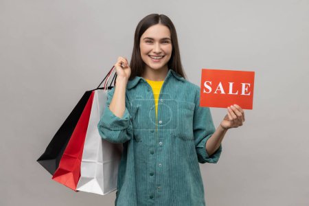 Photo for Friendly joyful woman with dark hair holding sale card and shopping bags, being happy with low prices, wearing casual style jacket. Indoor studio shot isolated on gray background. - Royalty Free Image
