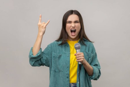 Photo for Portrait of attractive woman with dark hair singing loud favorite songs with microphone and showing rock and roll gesture, wearing casual style jacket. Indoor studio shot isolated on gray background. - Royalty Free Image