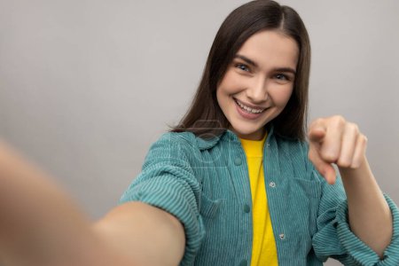 Photo for Woman with satisfied face expression, making selfie, expresses sincere emotions, pointing finger to camera, POV, wearing casual style jacket. Indoor studio shot isolated on gray background. - Royalty Free Image