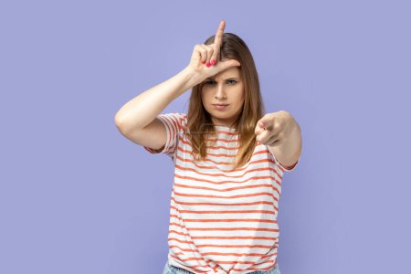 Photo for You are loser. Portrait of serious blond woman wearing striped T-shirt showing loser gesture and pointing finger on you, abuser. Indoor studio shot isolated on purple background. - Royalty Free Image