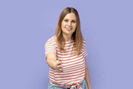Photo for Let me introduce myself. Portrait of friendly blond woman wearing striped T-shirt giving hand to handshake, greeting guests with toothy smile. Indoor studio shot isolated on purple background. - Royalty Free Image