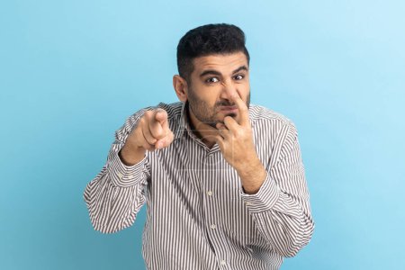 Photo for You are liar. Portrait of angry bearded man touching his nose with finger and showing lie gesture, body language, wearing striped shirt. Indoor studio shot isolated on blue background. - Royalty Free Image