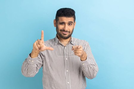 Photo for Portrait of disrespectful rude young businessman showing loser gesture and pointing on you, abuser, frowning face, wearing striped shirt. Indoor studio shot isolated on blue background. - Royalty Free Image