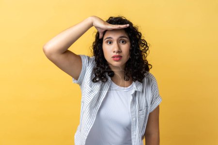 Photo for Portrait of attractive young adult woman with dark wavy hair keeps hand near forehead looks far away searches something on horizon. Indoor studio shot isolated on yellow background. - Royalty Free Image