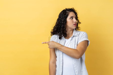 Photo for Portrait of woman with dark wavy hair pointing finger aside, ordering get out and looking resentful, boss dismissing from work, showing exit. Indoor studio shot isolated on yellow background. - Royalty Free Image