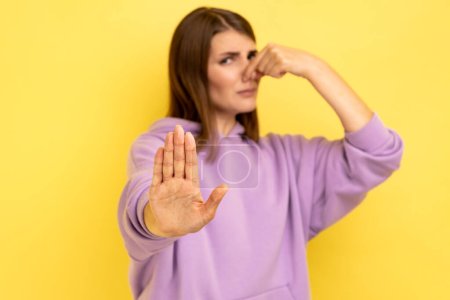 Portrait of woman grimacing with disgust, holding breath, pinching nose with fingers to avoid bad smell, showing stop gesture, wearing purple hoodie. Indoor studio shot isolated on yellow background