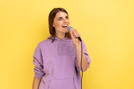 Téléchargez les photos : Portrait de femme positive chantant bruyamment, tenant le microphone à la main, s'amusant à se reposer dans le karaoké, chanteuse, portant un sweat à capuche violet. Studio intérieur tourné isolé sur fond jaune. - en image libre de droit