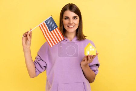 Photo for Real estate purchase in America, immigration. Portrait of happy woman holding american flag and paper house, wearing purple hoodie. Indoor studio shot isolated on yellow background. - Royalty Free Image
