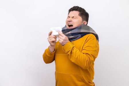 Photo for Man wrapped in scarf sneezing in napkin, cleaning running nose and coughing, suffering influenza symptoms, fever, wearing urban style hoodie. Indoor studio shot isolated on white background. - Royalty Free Image
