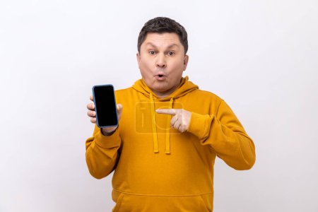 Photo for Man pointing finger at smartphone with empty screen, looking at camera with shocked expression, freespace for advertisement, wearing urban style hoodie. Indoor studio shot isolated on white background - Royalty Free Image