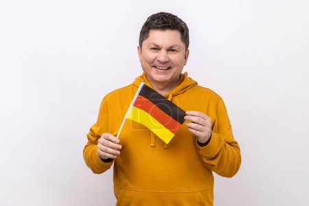 Téléchargez les photos : Satisfait homme d'âge moyen positif tenant le drapeau allemand dans sa main et regardant la caméra avec un sourire de dents, portant un sweat à capuche de style urbain. Studio intérieur tourné isolé sur fond blanc. - en image libre de droit