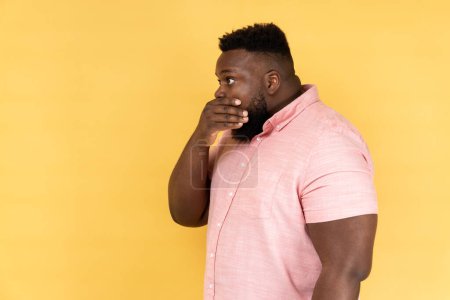 Photo for I won't tell. Side view of handsome man wearing pink shirt covering mouth with hand, keeping secret, terrified with shocking news. Indoor studio shot isolated on yellow background. - Royalty Free Image