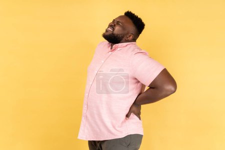 Photo for Back, kidney or spine pain. Portrait of man wearing pink shirt standing and endure pain on back, frowning face, suffering terrible pain. Indoor studio shot isolated on yellow background. - Royalty Free Image