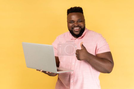 Téléchargez les photos : Portrait d'un homme souriant tenant un ordinateur portable à la main montrant les pouces vers le haut geste, blogueur aime les messages, regarder la caméra, recommander aux abonnés. Studio intérieur tourné isolé sur fond jaune. - en image libre de droit