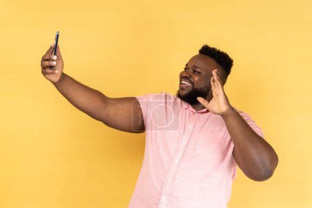 Téléchargez les photos : Portrait d'un homme barbu portant une chemise rose disant bonjour en faisant un appel vidéo à l'aide d'un smartphone, souriant avec le sourire des dents, connexion 4g. Studio intérieur tourné isolé sur fond jaune. - en image libre de droit