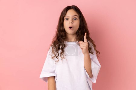 Porträt eines überraschten kleinen Mädchens in weißem T-Shirt, das den Finger in die Höhe reckt, hat plötzlich eine großartige Idee und blickt mit offenem Mund in die Kamera. Indoor-Studio isoliert auf rosa Hintergrund aufgenommen.