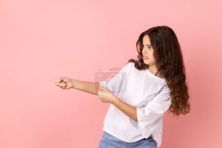 Photo for Side view portrait of adorable charming little girl wearing white T-shirt standing in attack or pulling hands gesture and looking forward. Indoor studio shot isolated on pink background. - Royalty Free Image