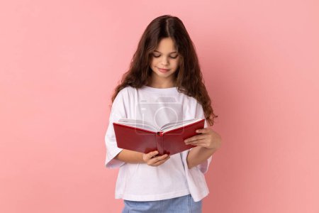 Photo for Portrait of delighted charming little girl wearing white T-shirt holding in hands and reading interesting book, being very attentive. Indoor studio shot isolated on pink background. - Royalty Free Image