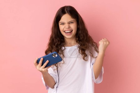 Foto de Retrato de una niña pequeña con una camiseta blanca de pie, usando un teléfono inteligente y jugando al juego móvil con la cara positiva emocionada, el puño cerrado. Estudio interior plano aislado sobre fondo rosa. - Imagen libre de derechos