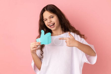 Photo for Portrait of happy satisfied adorable little girl wearing white T-shirt holding and pointing at like or thumbs up paper shape sign. Indoor studio shot isolated on pink background. - Royalty Free Image
