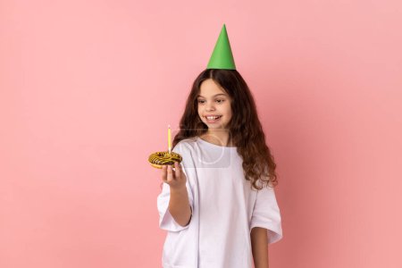 Téléchargez les photos : Petite fille satisfaite en cône de fête vert sur la tête tenant gâteau avec bougie et regardant le dessert avec sourire dents, exprimant le pur bonheur de l'enfant. Studio intérieur tourné isolé sur fond rose. - en image libre de droit
