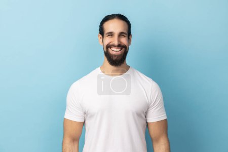 Foto de Retrato de un hombre guapo barbudo con una camiseta blanca de pie mirando a la cámara con la cara satisfecha y sonriendo, expresando felicidad. Estudio interior plano aislado sobre fondo azul. - Imagen libre de derechos