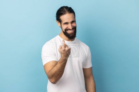 Foto de Retrato de un hombre positivo con barba usando una camiseta blanca haciendo un gesto de señas, llamando invitando a acercarse, sonriendo y coqueteando. Estudio interior plano aislado sobre fondo azul. - Imagen libre de derechos