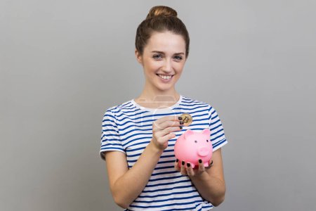 Téléchargez les photos : Portrait de femme portant T-shirt rayé mettant Bitcoin physique dans la tirelire, investissement dans le concept de crypto-monnaie, regardant la caméra en souriant. Studio intérieur tourné isolé sur fond gris. - en image libre de droit