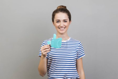 Photo for Portrait of woman wearing striped T-shirt showing thumbs up blue paper sign, liking and recommending posts in social networks, asking to rate. Indoor studio shot isolated on gray background. - Royalty Free Image