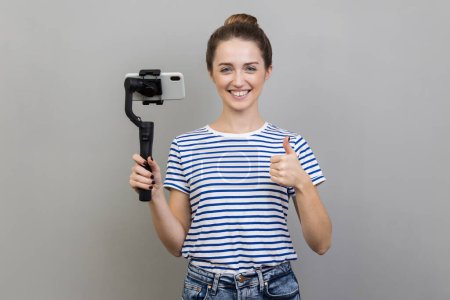 Photo for Satisfied stylish woman wearing striped T-shirt holding mobile phone and steadicam, looking at camera with positive expression, showing thumb up. Indoor studio shot isolated on gray background. - Royalty Free Image