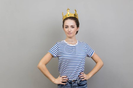 Photo for Portrait of woman wearing striped T-shirt and golden crown, looking at camera with confident facial expression, keeps hands on hips. Indoor studio shot isolated on gray background. - Royalty Free Image