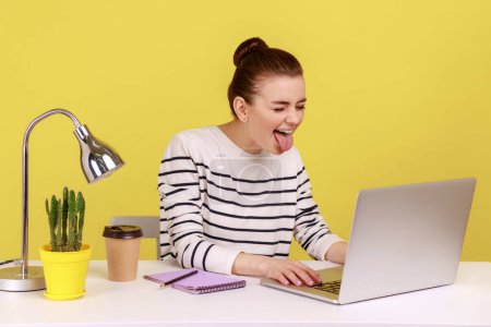 Photo for Playful funny woman office worker in striped shirt showing tongue out, expressing positive childish emotions, chatting via web camera on laptop. Indoor studio studio shot isolated on yellow background - Royalty Free Image