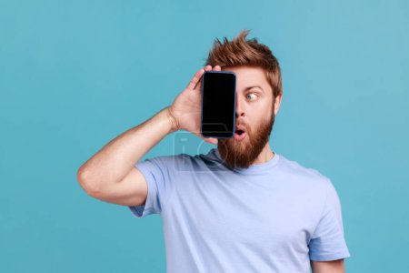 Téléchargez les photos : Portrait d'un homme barbu couvrant l'œil avec un téléphone portable, cachant la moitié du visage, utilisant un gadget avec une expression choquée, essayant de regarder l'écran. Studio intérieur tourné isolé sur fond bleu. - en image libre de droit
