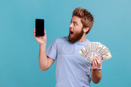Photo for Portrait of surprised amazed handsome bearded man holding dollar banknotes and looking at mobile phone with empty screen, online betting, winning. Indoor studio shot isolated on blue background. - Royalty Free Image