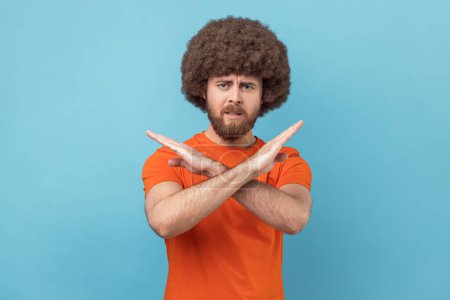 Photo for Portrait of man with Afro hairstyle wearing orange T-shirt showing cross, making stop gesture, saying no with angry expression, declines something. Indoor studio shot isolated on blue background. - Royalty Free Image