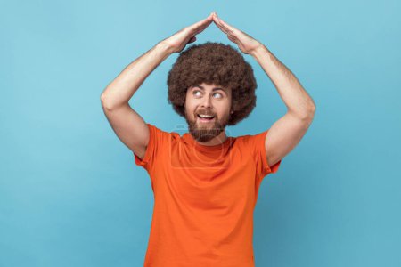 Photo for Portrait of man with Afro hairstyle wearing orange T-shirt standing raising hands showing roof gesture and smiling contentedly, dreaming of house. Indoor studio shot isolated on blue background. - Royalty Free Image