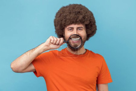 Téléchargez les photos : Portrait d'homme drôle avec une coiffure afro portant un T-shirt orange debout, tenant une loupe sur ses dents, regardant avec un sourire de dents. Studio intérieur tourné isolé sur fond bleu. - en image libre de droit