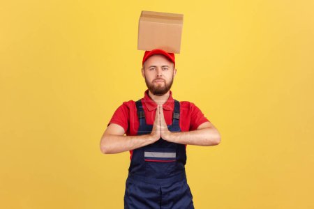 Photo for Relaxed courier man standing with cardboard box on his head, keeping hands in pray gesture, trying to calm down and resting during working day. Indoor studio shot isolated on yellow background. - Royalty Free Image