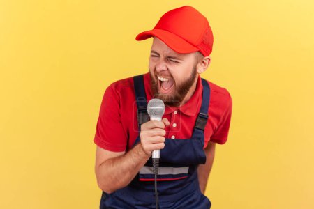 Photo for Portrait of extremely excited worker man wearing blue uniform and red cap relaxing at a corporate party singing karaoke at work. Indoor studio shot isolated on yellow background. - Royalty Free Image