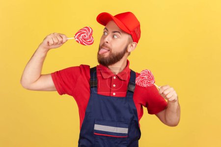 Photo for Portrait of funny hungry worker man wearing red cap and blue uniform holding tasty sugary candy, licking heart shape lollypop, tongue out. Indoor studio shot isolated on yellow background. - Royalty Free Image