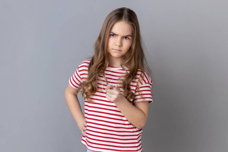 Photo for Portrait of little girl wearing striped T-shirt pointing finger at camera and looking with dissatisfied suspicious expression, warning about troubles. Indoor studio shot isolated on gray background. - Royalty Free Image