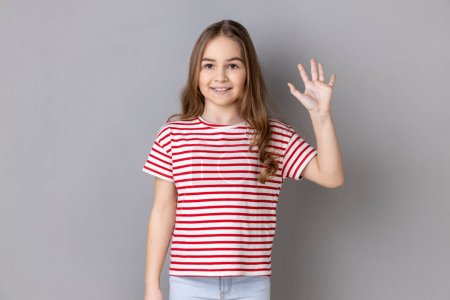Retrato de una adorable niña alegre con una camiseta a rayas de pie saludando con la mano, mirando a la cámara con una sonrisa dentada atractiva. Estudio interior plano aislado sobre fondo gris.