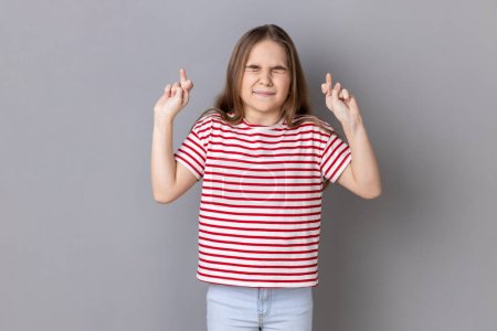 Photo for Portrait of little girl wearing striped T-shirt holding crossed fingers and wishing for good luck, having hopeful look, hoping for victory. Indoor studio shot isolated on gray background. - Royalty Free Image