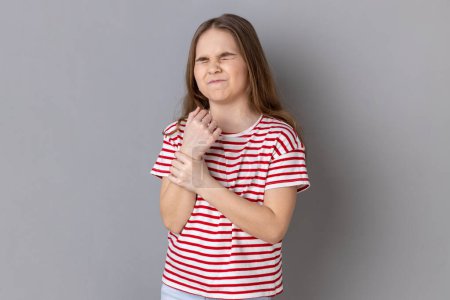 Photo for Portrait of sick little girl wearing striped T-shirt standing with grimace of pain, massaging sore wrist, suffering hand injury or sprain. Indoor studio shot isolated on gray background. - Royalty Free Image