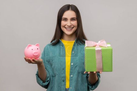 Photo for Portrait of happy woman holding wrapped gift box and piggy bank, save money, advertising, looking smiling at camera, wearing casual style jacket. Indoor studio shot isolated on gray background. - Royalty Free Image