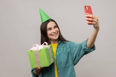 Photo for Portrait of smiling woman holding present box in hands and taking selfie or having video call with client, wearing casual style jacket. Indoor studio shot isolated on gray background. - Royalty Free Image